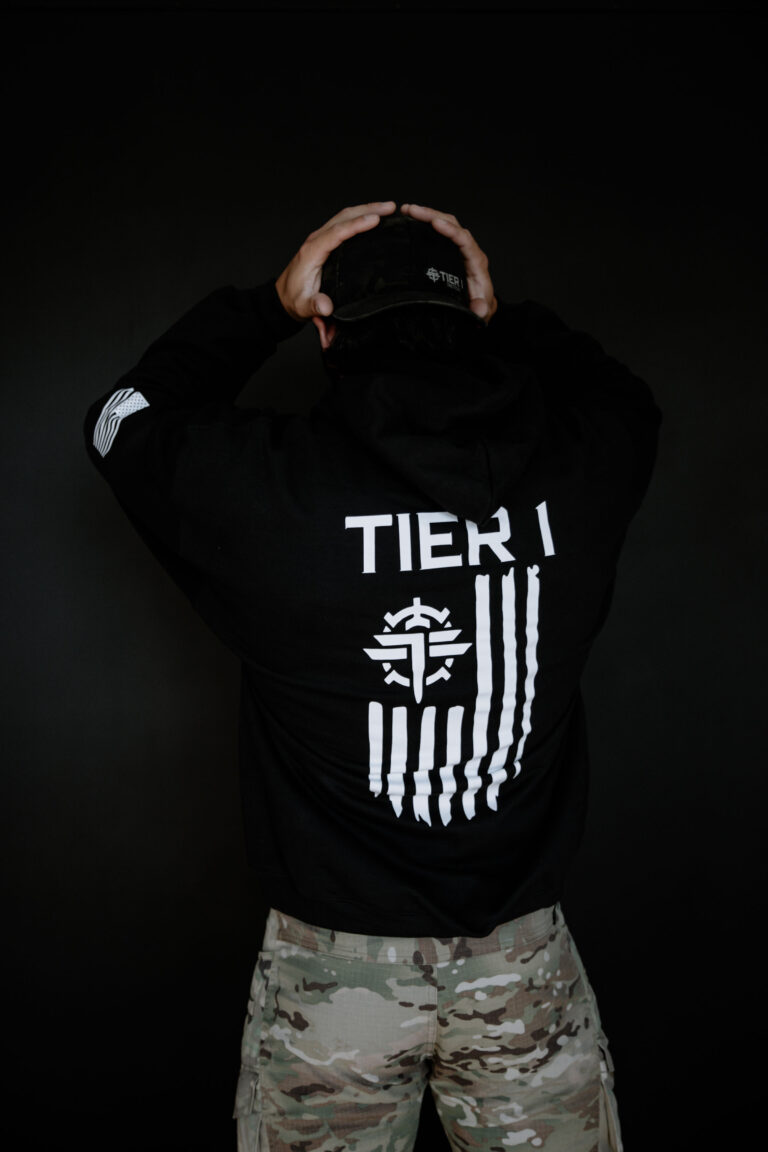 Back view of a person in a black hoodie with 'TIER 1' graphic on the back, hands on a camouflage cap, dark background.