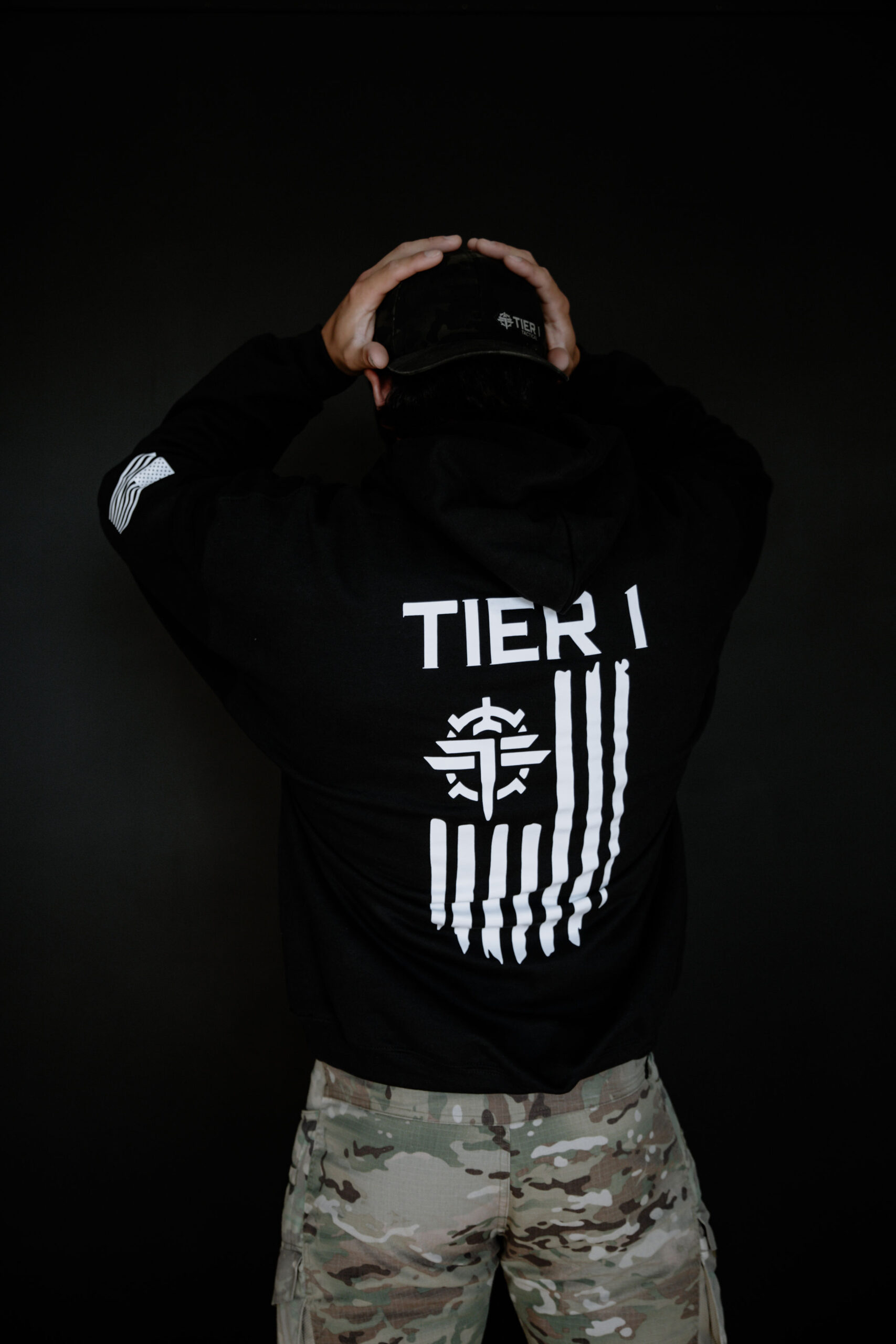 Back view of a person in a black hoodie with 'TIER 1' graphic on the back, hands on a camouflage cap, dark background.