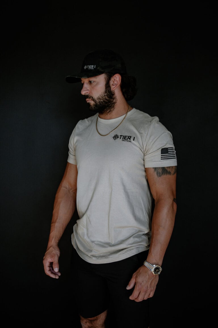 Bearded man in a beige Tier 1 Tactical T-shirt, camo cap, and gold chain, facing left against a dark background.